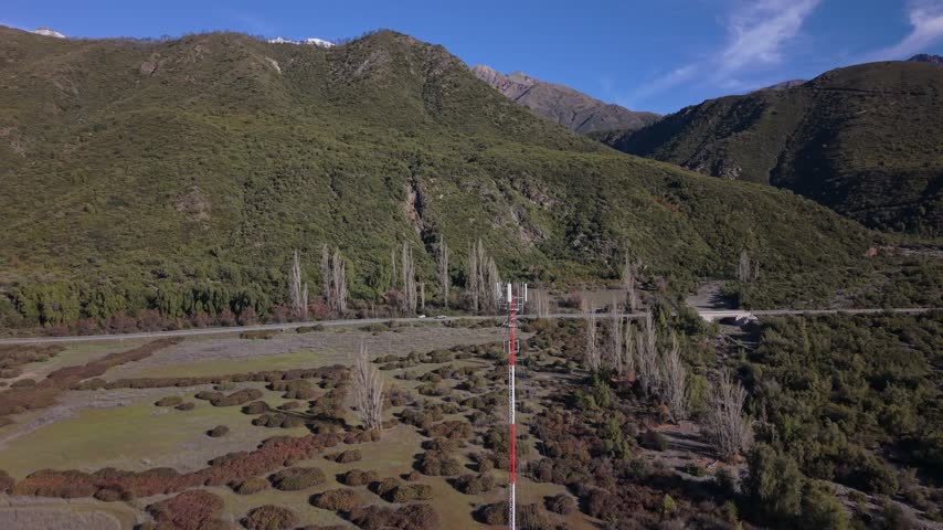 Ascending tilt down shot focusing on a slender telco tower on a shrubland valley floor beneath a forested mountain slope near a rural road