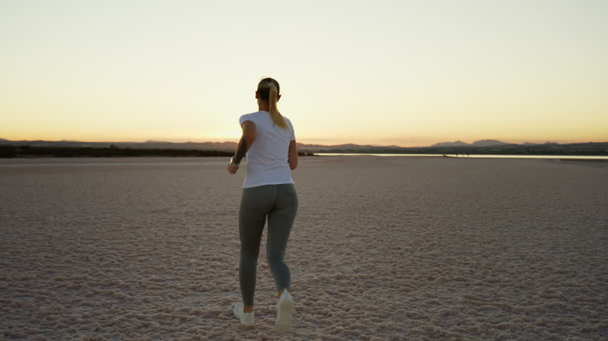 Young athletic woman running away from the camera in a surreal, expansive salt flat landscape during a beautiful sunset, embodying a healthy lifestyle, determination, and freedom