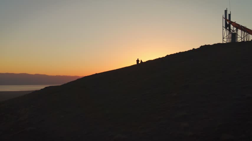 Aerial silhouette of hikers standing on mountain pass near communication tower watching golden sunset over Song-Kul Lake, Kyrgyzstan