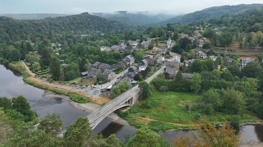 Picturesque landscape featuring a winding river flowing past a rural village with stone buildings and a bridge under a soft overcast sky in France