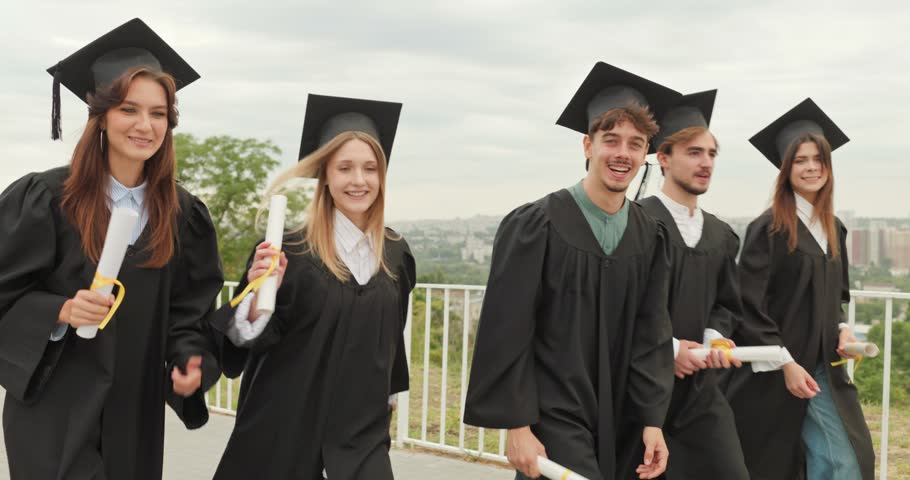 Graduate friends celebrate outdoors with diplomas and smiles. Young students in caps and gowns laugh and dance toward camera during ceremony. A very joyful moment of shared achievement.