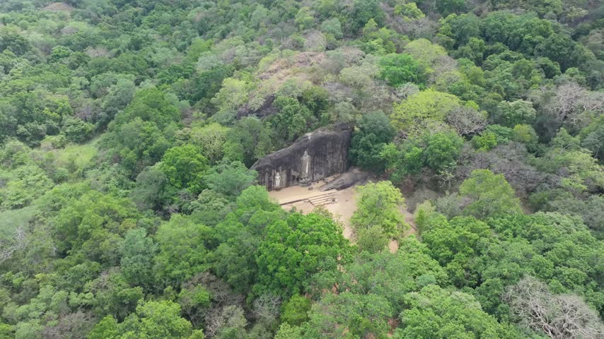 Aerial view of an ancient rock temple carved into a large boulder surrounded by dense tropical forest in Ambagollapathana, Uva Province, Sri Lanka.