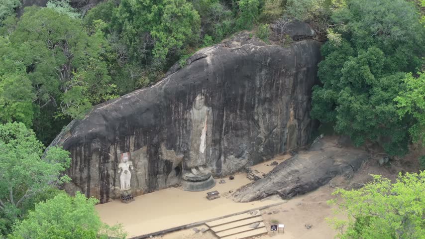 Aerial view of the ancient Buduruvagala rock carvings featuring large Buddha statues carved into a cliff face surrounded by lush forest in Ambagollapathana, Uva Province, Sri Lanka.