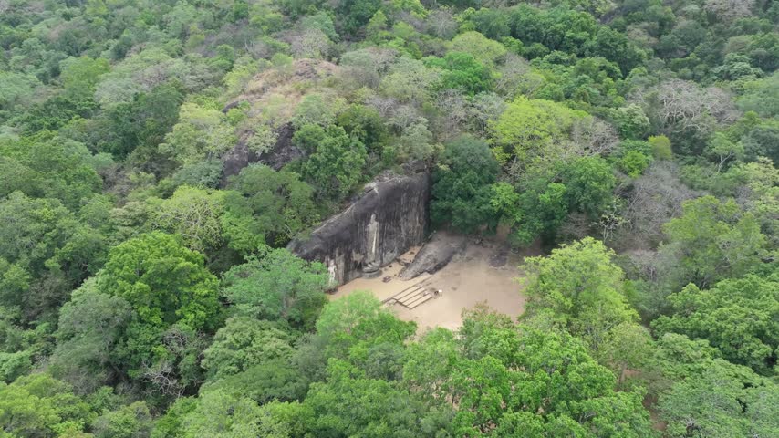 Aerial view of an ancient rock-cut Buddha statue carved into a large cliff face surrounded by dense tropical forest in Ambagollapathana, Uva Province, Sri Lanka.
