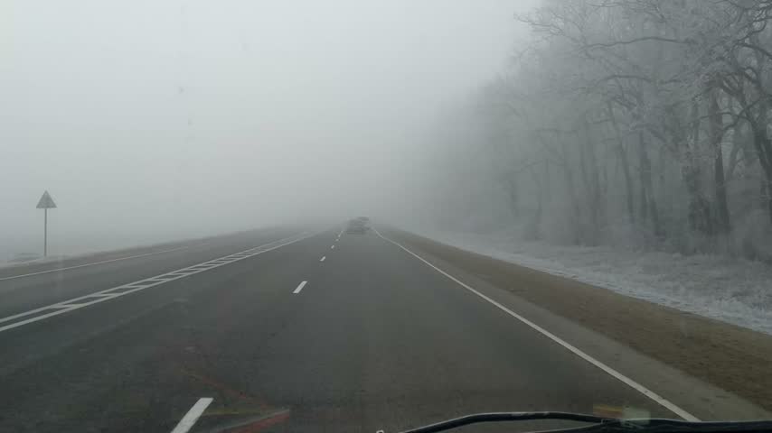 Point of view driving along an asphalt highway through thick white fog past frost covered trees in winter, poor road visibility.