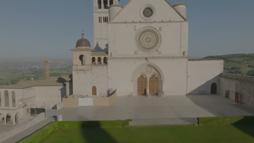 Aerial view of the Basilica of Saint Francis with its iconic rose window and stone facade set against the green hills of Assisi, Umbria, Italy.
