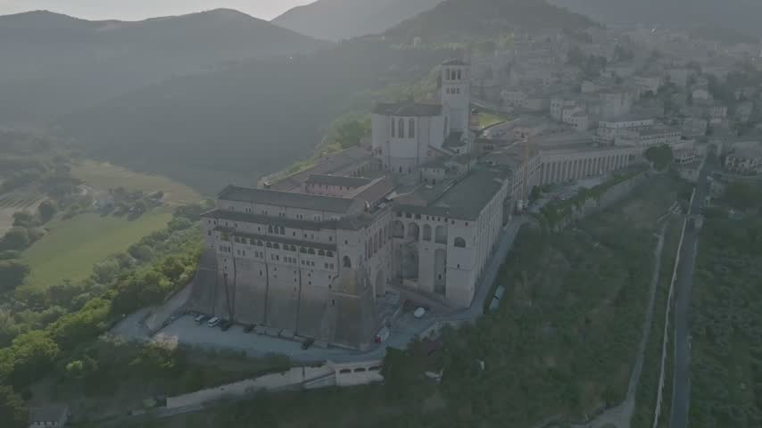Aerial view of the historic Basilica of Saint Francis of Assisi perched on a hilltop surrounded by lush green hills in Assisi, Umbria, Italy.