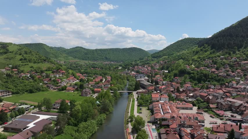 Aerial view of the town of Visoko with a river flowing through the center, surrounded by lush green hills and residential houses with red roofs in Visoko, Federation of Bosnia, Bosnia and Herzegovina.