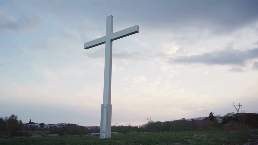 Large white christian cross standing on green hill with cloudy sky during sunset in serene landscape scene.