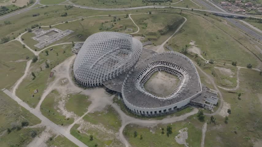 Aerial view of the unfinished Vela di Calatrava sports complex with its distinctive white steel structure and concrete stadium bowl surrounded by green fields in Roma, Lazio, Italy.