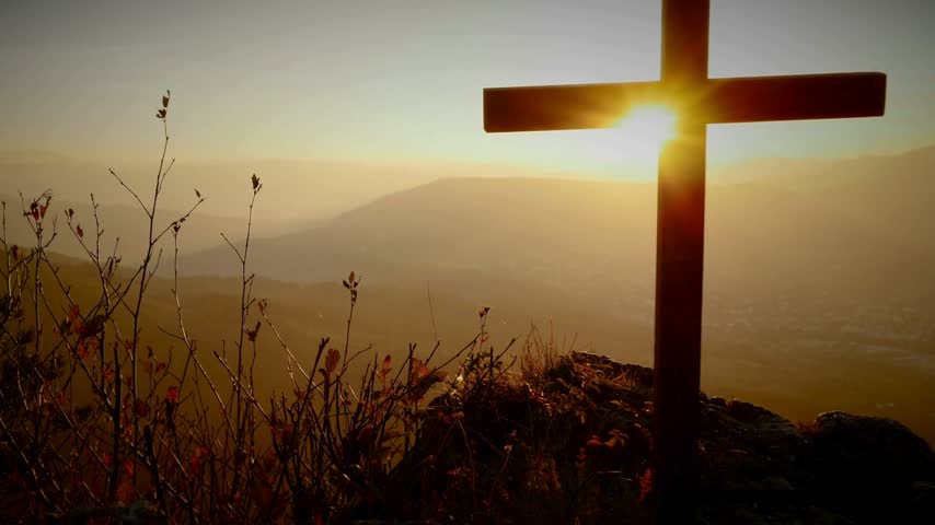 Sunlight rays glowing behind wooden cross silhouette on hilltop with panoramic mountain valley landscape.