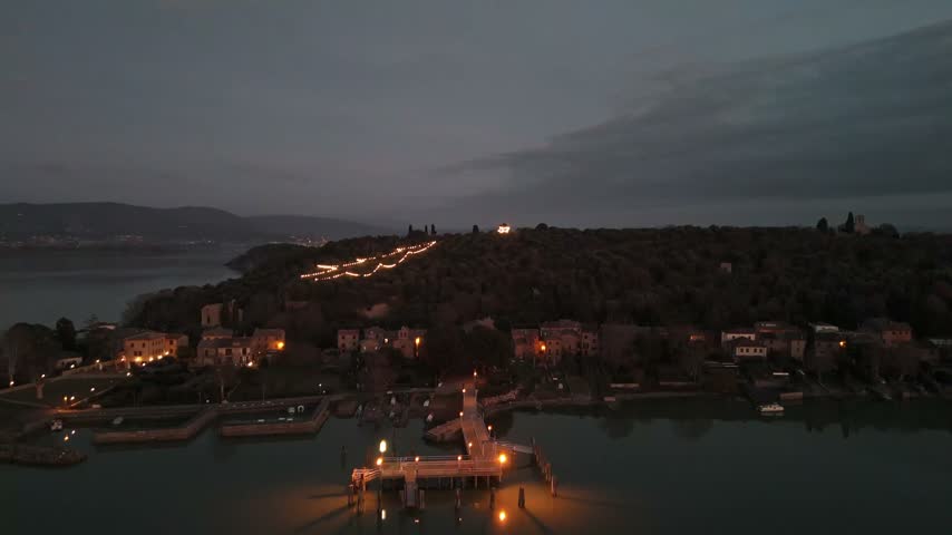 Aerial view of the illuminated pier and historic village of Isola Maggiore on Lake Trasimeno at dusk in Tuoro sul Trasimeno, Umbria, Italy.