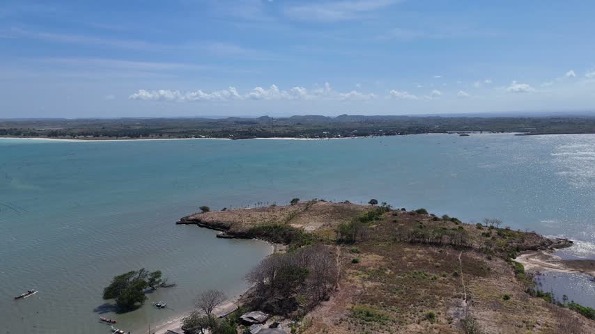 Aerial view of a tranquil bay with dry coastal land, scattered trees, small fishing boats, and blue ocean waters under a clear sky.
