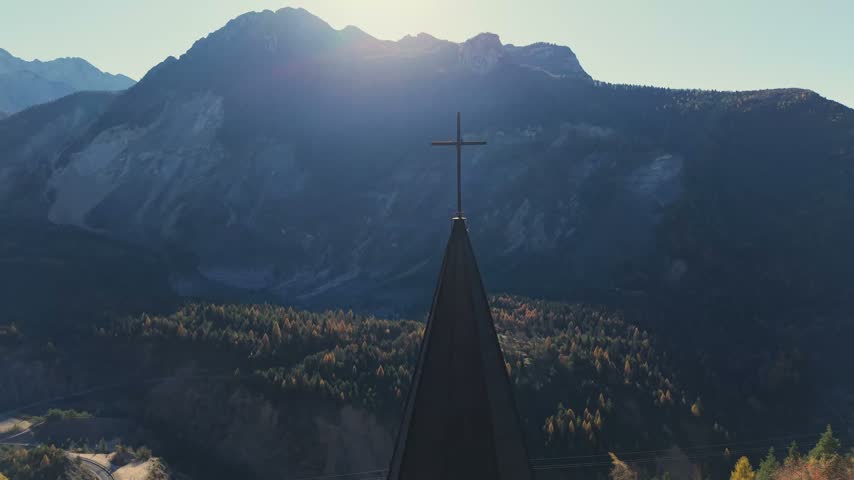 Aerial view of a church steeple with a cross set against a backdrop of mountains and forest in the golden hour light in Pordenone, Friuli-Venezia Giulia, Italy.
