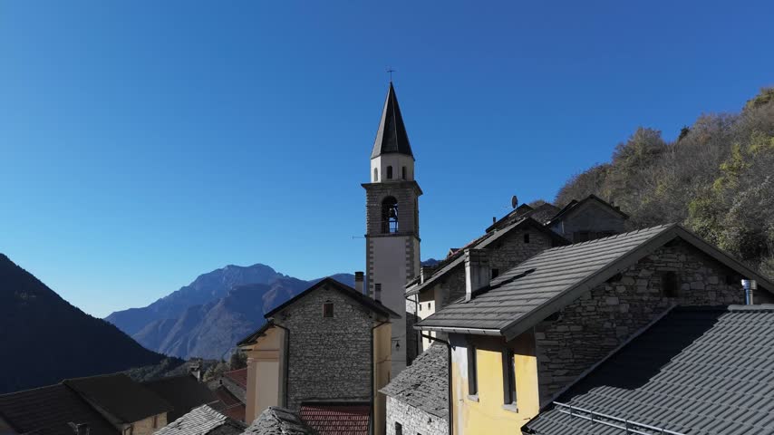 Aerial view of a historic church tower rising above stone houses in a mountain village under a clear blue sky in Pordenone, Friuli-Venezia Giulia, Italy.