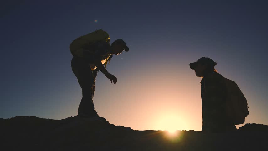 Business team silhouette climb. A man helps a woman climb a rocky surface as the sun sets behind them. The man extends his hand to assist the woman during their hike.