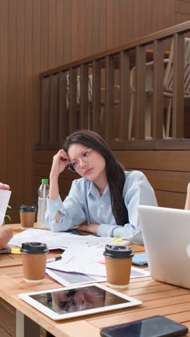Exhausted businesswoman looks stressed while colleagues discuss work around a cluttered conference table.