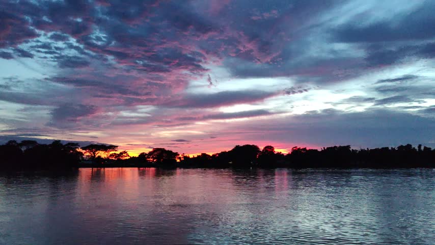 Serene sunset landscape with a calm lake, dramatic clouds, dark tree-lined horizon, distant bridge, and vivid orange reflections shimmering across the water.