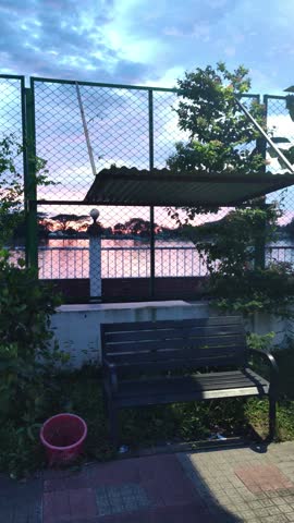 Twilight lakeside park with a dark slatted bench, red bucket, chain-link fence, colorful sunset reflections, and a glowing street lamp over a tiled walkway.