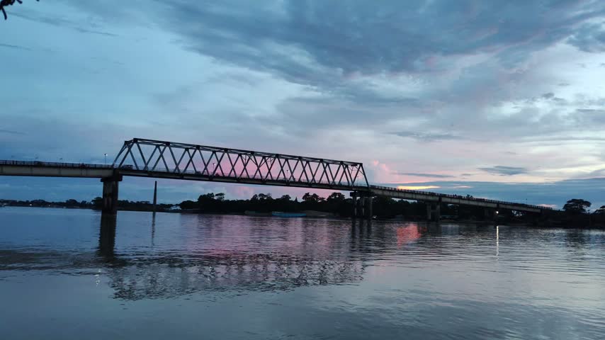 Serene twilight river scene with a dramatic truss bridge, soft pink-purple sky, calm reflections, weathered railing, and barbed wire framing the landscape.