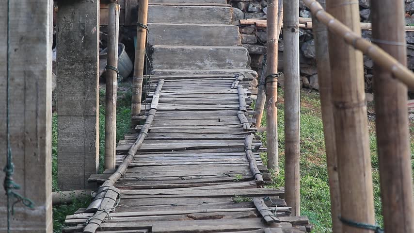 Narrow wooden walkway between tall bamboo poles leading forward through lush green foliage and soft misty atmosphere