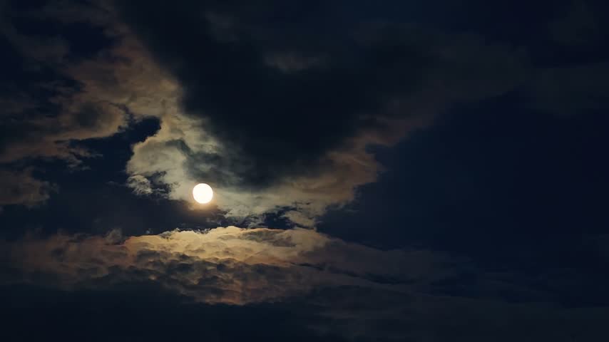 A high-angle video capturing a bright full moon glowing vividly as it peaks through thick, dark dramatic storm clouds, creating a mysterious, eerie, and atmospheric nighttime sky view during a cloudy lunar evening.
