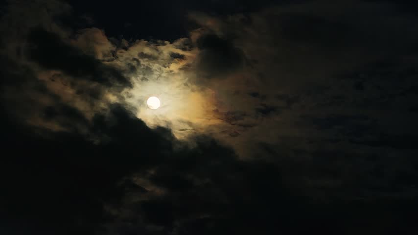 A high-angle video capturing a bright full moon glowing vividly as it peaks through thick, dark dramatic storm clouds, creating a mysterious, eerie, and atmospheric nighttime sky view during a cloudy lunar evening.