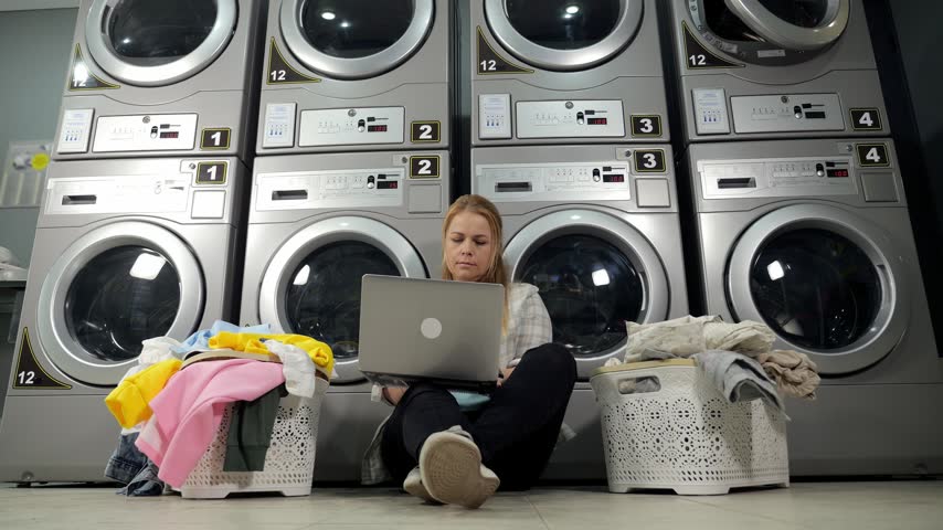 A woman with a laptop is sitting on the laundry room floor against the background of a row of washing machines and dryers, next to baskets of dirty laundry. The woman is waiting for the end of washing