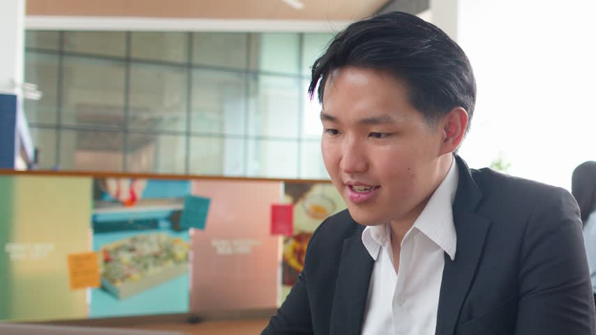 An Asian businessman in a suit types on his laptop in a bright coworking space.