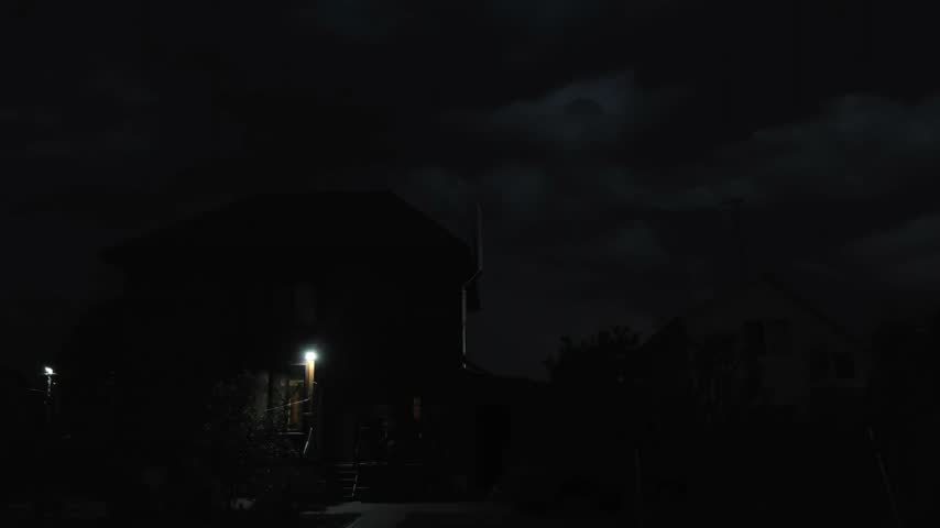 Residential houses under storm clouds as evening fades into night, with a porch light illuminating a quiet suburban yard