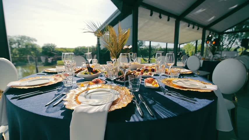 Beautifully decorated elegant banquet table set with a blue tablecloth, gold plates, and appetizers for a formal event at an outdoor venue with a scenic lake view