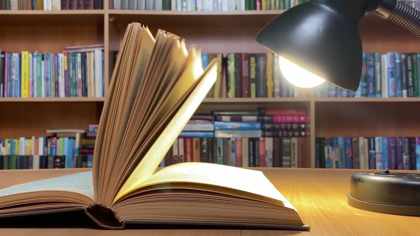 The wind gently blows through the pages of a thick open book lying on a wooden desk illuminated by a glowing lamp in a quiet library