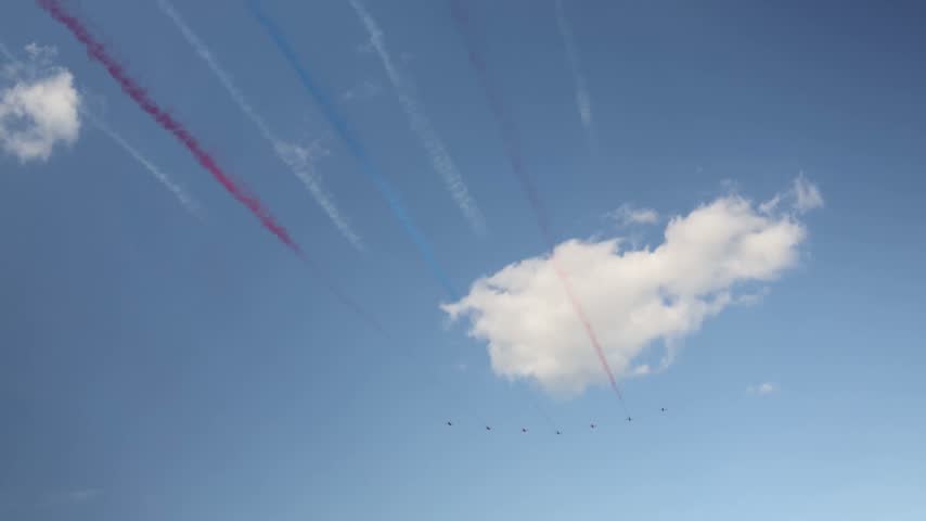 Aerobatic jet formation flies across blue sky leaving red white and blue smoke trails during an airshow creating a dramatic aviation display high above the crowd below us