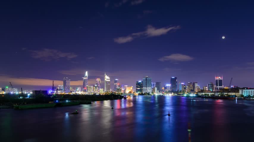 Nighttime waterfront cityscape with illuminated buildings reflecting across the calm water under a dramatic evening sky.