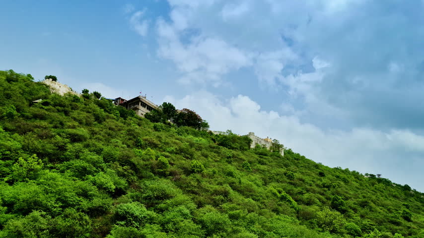 A broad green hillside slopes beneath hilltop structures and scattered white clouds in Udaipur.