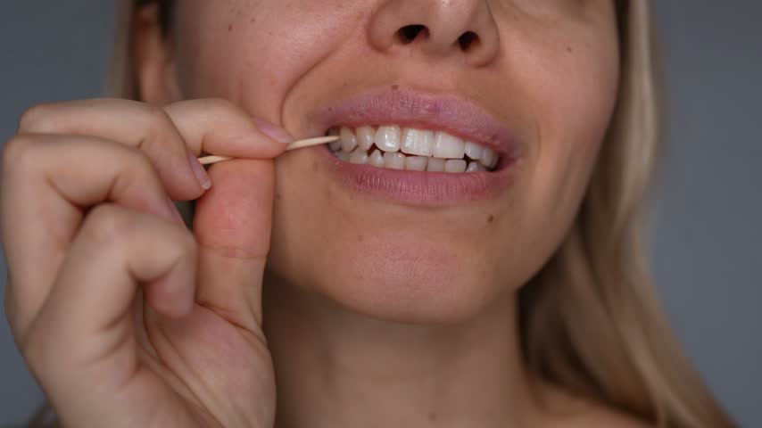 Young woman using a wooden toothpick to clean food between teeth, close-up of mouth, lips and hand during dental hygiene routine on gray background