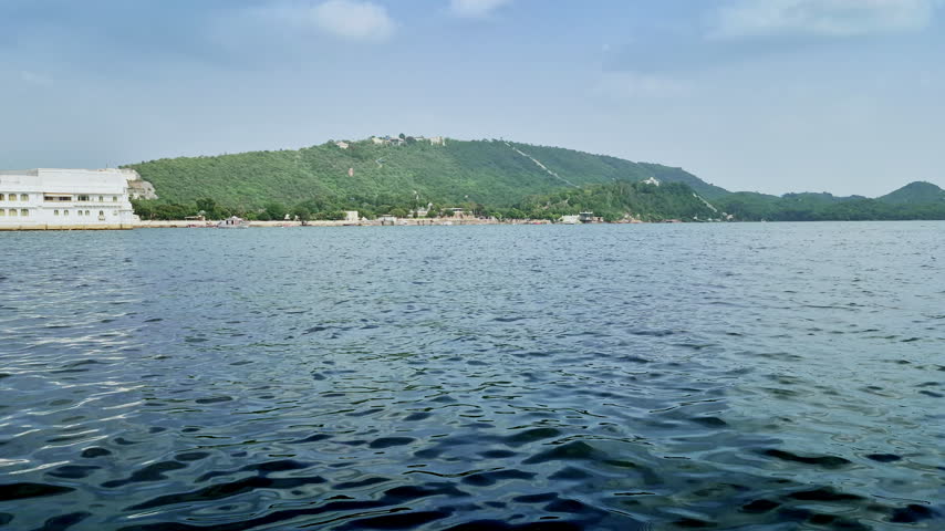 A green hill with pale structures and a descending hillside wall rises beyond Lake Pichola in Udaipur.