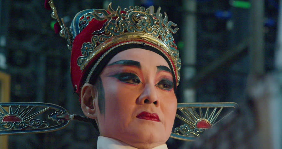 A Chinese opera actress in an ornate crown and orange costume sits backstage, ready to perform.