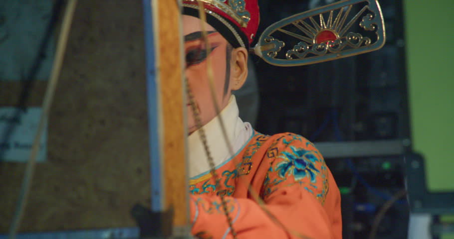 A Chinese opera actress in an ornate crown and orange costume sits backstage, ready to perform.