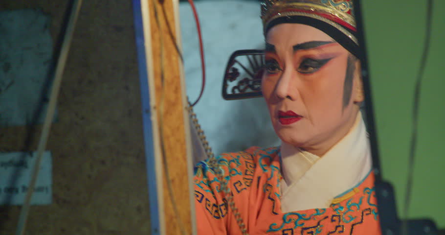 A Chinese opera actress in an ornate crown and orange costume sits backstage, ready to perform.