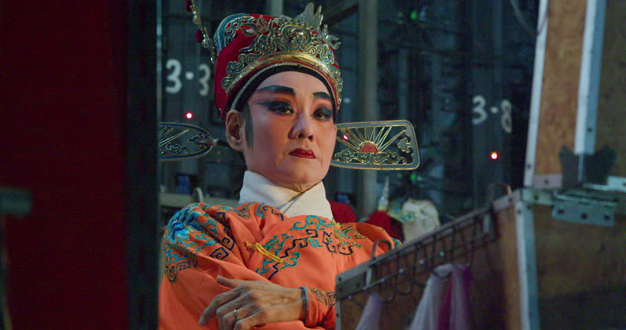 A Chinese opera actress in an ornate crown and orange costume sits backstage, ready to perform.