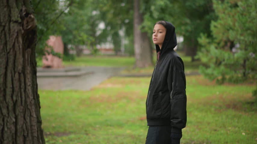 Female in rain gazes thoughtfully. Lady in dark hoodie pauses to reflect amidst greenery and rain. Woman in black hoodie stands in rainfilled park gazing at trees and reflecting quietly