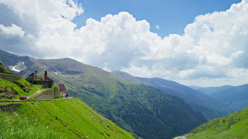 Windswept terrain presents Transfagarasan crossing steep green Capra Valley slopes, near remote mountain buildings.