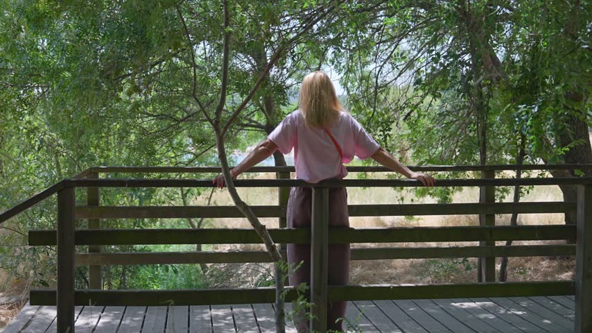 Young woman from behind standing on a wooden walkway in a forest. Solo female traveler enjoying the tranquil scenery of a green park