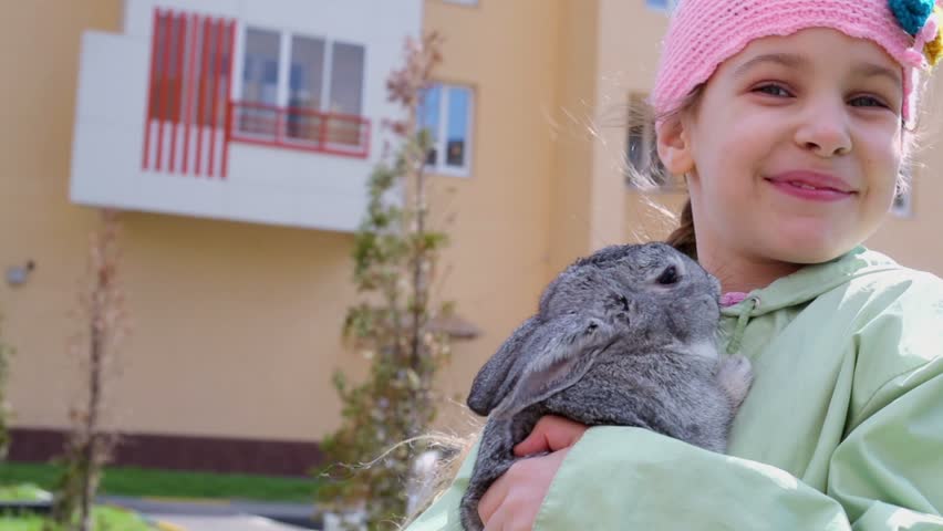 Little girl in greenish jacket and knitted hat holds rabbit in her hands near dwelling house