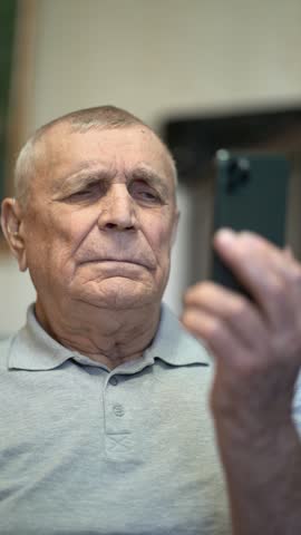 Close-up of elderly senior man holding phone and viewing screen content indoors in casual polo shirt.