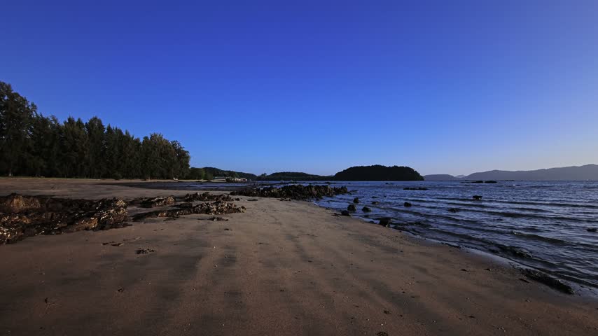 An empty sandy beach on a tropical island has exposed rocks and shallow sea water at low tide. A rocky shoreline meets the calm sea, with tropical forest on the coast and distant islands across the bay. Wet sand, coastal rocks and small waves fill this deserted beach. This coastline has a wide sandy shore, ocean view and mountains on the horizon. No people are visible on this remote tropical beach in Thailand, Southeast Asia.