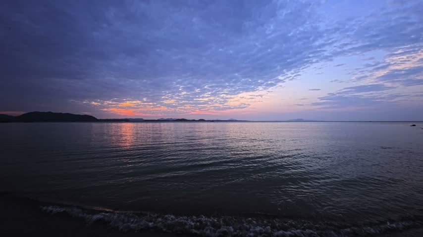 
Sunrise over a calm tropical sea, with orange morning light reflecting on the water beneath a wide blue cloudy sky. Distant islands and low mountains stretch along the ocean horizon as small waves ripple across the foreground. Pink and orange colors appear between clouds above the island silhouettes. This sunrise seascape shows a tropical island, calm water, cloud cover, dawn sky, sea reflection and an open coastal view in Thailand, Southeast Asia.