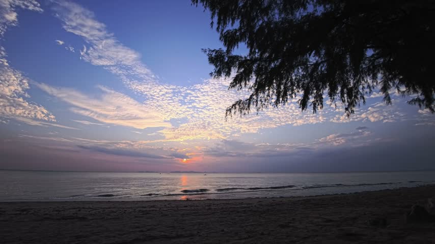 Sunset over a tropical beach with dark sand, calm sea and small waves reaching shore. The setting sun reflects on the water below a cloudy sky, with distant islands visible along the horizon. Overhanging coastal trees frame this beach while orange and blue evening light spreads across the ocean. An empty sandy coast, sunset beach, sea view and tropical island at dusk in Thailand, Southeast Asia.