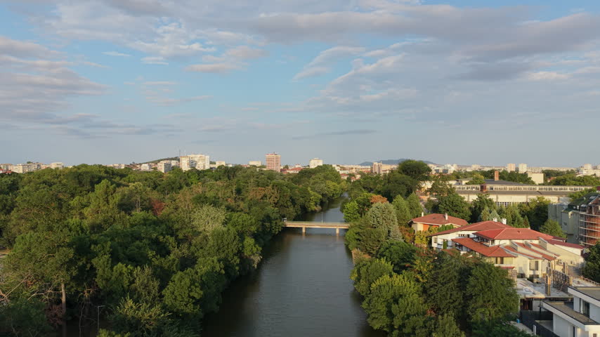 Aerial drone footage of the Tundzha River flowing through Yambol, Bulgaria, with a bridge, green riverside vegetation, residential buildings and the city skyline in the background